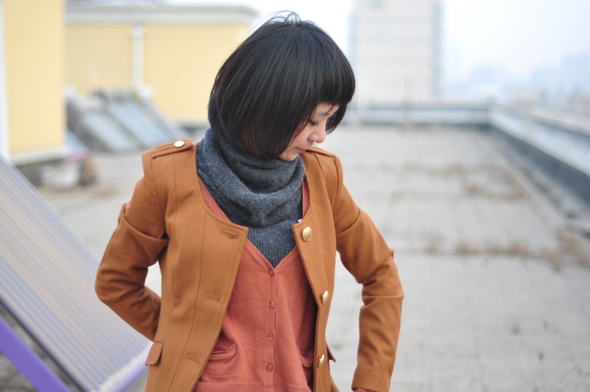 Woman in brown coat and scarf standing on rooftop with urban background.