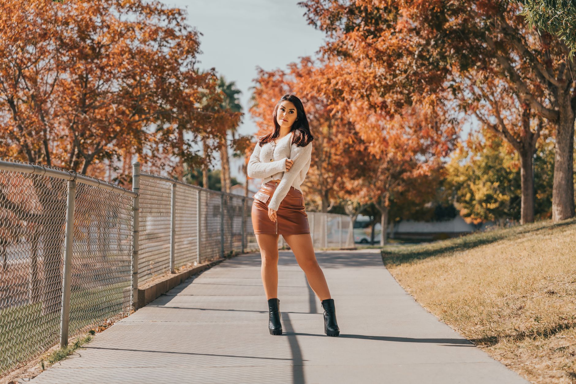 Stylish woman in white top and brown skirt standing confidently on a tree-lined street in autumn.