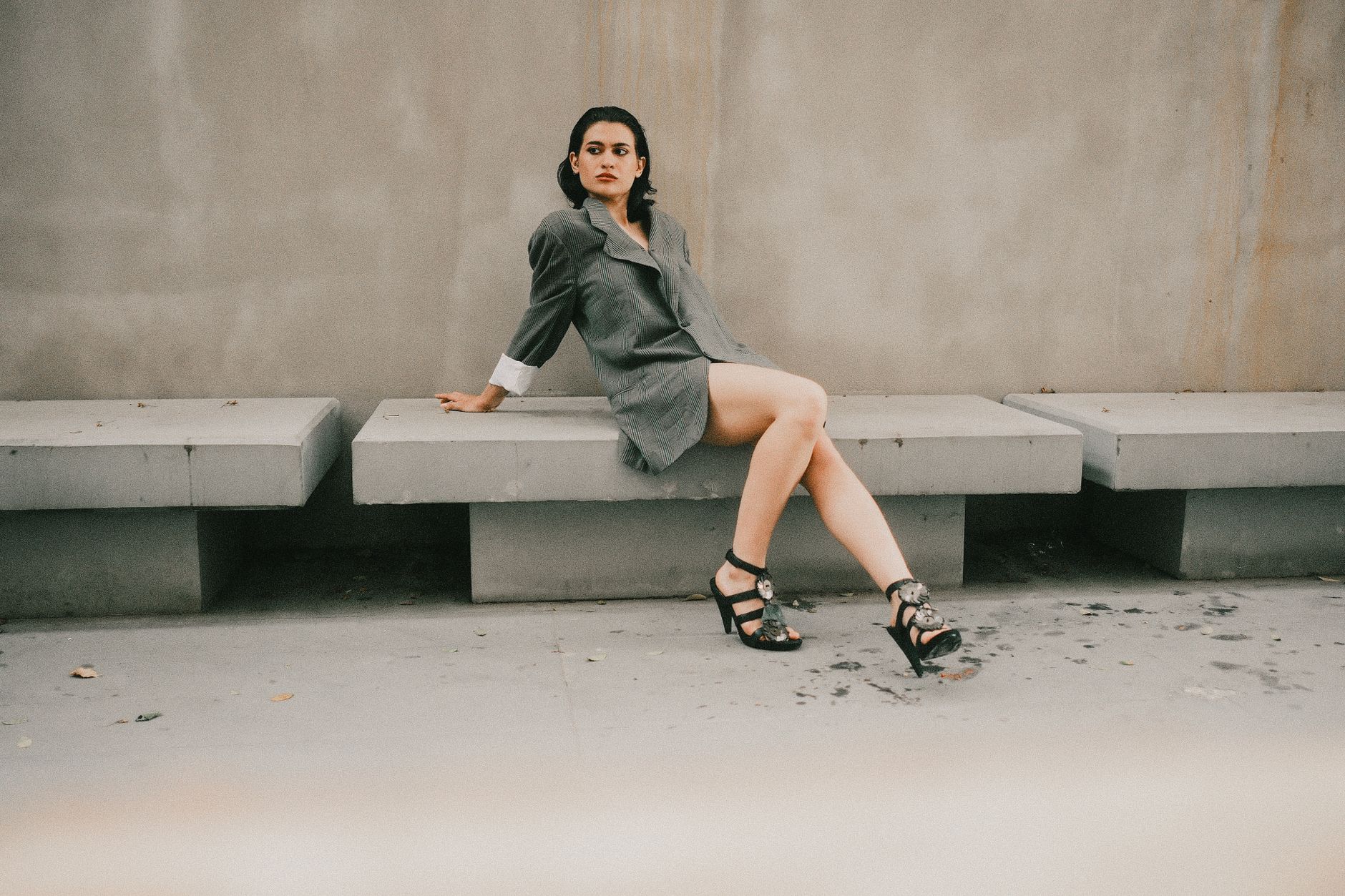 Elegant woman in gray blazer and heels poses on minimalist concrete bench outdoors.