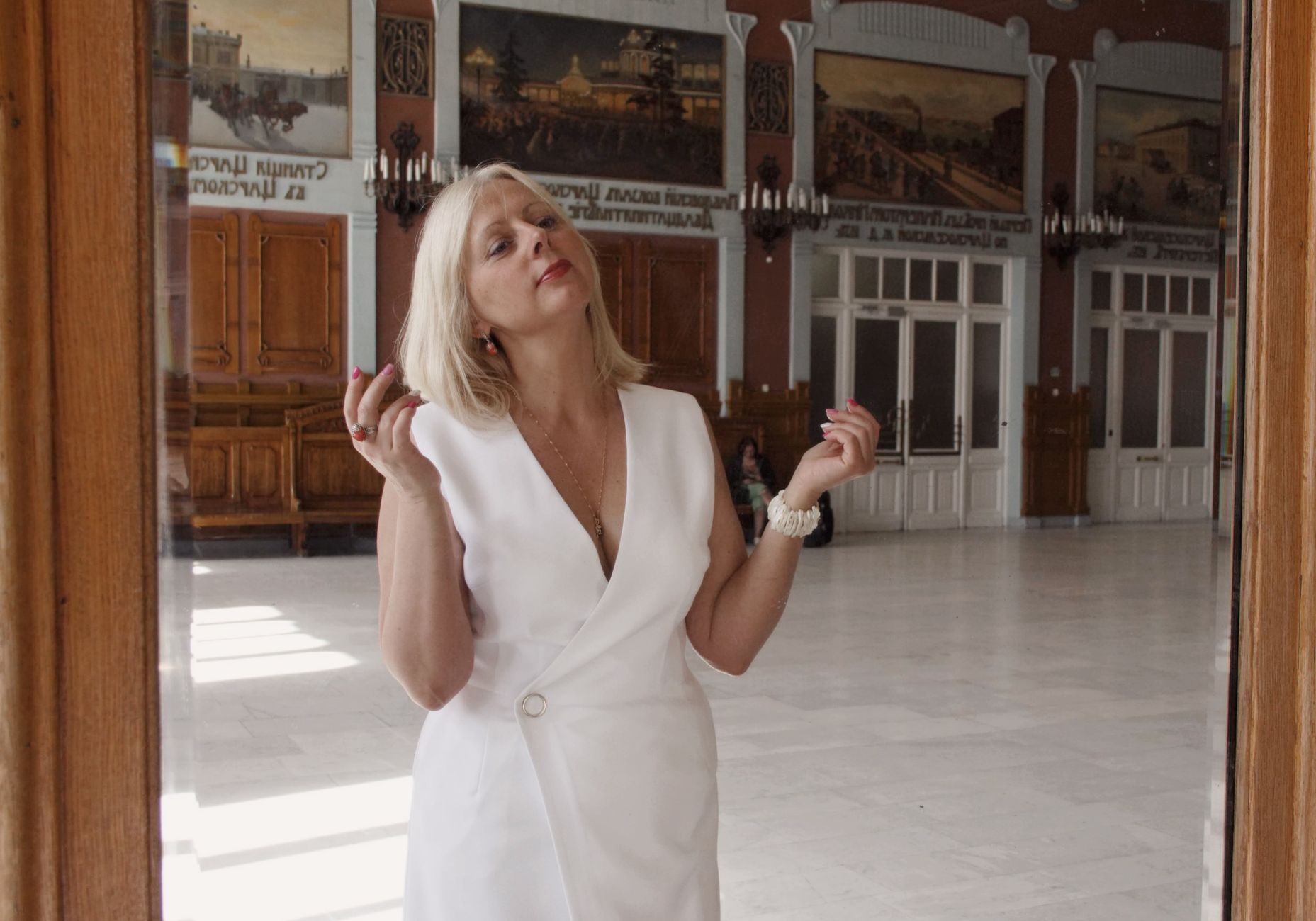 Blonde woman in white dress posing confidently in a historical indoor setting with ornate design.