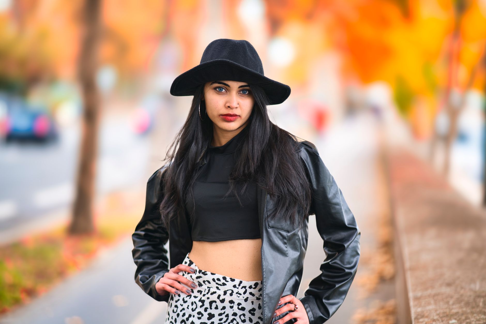 Stylish woman in black hat and leather jacket posing in urban area with autumn foliage.