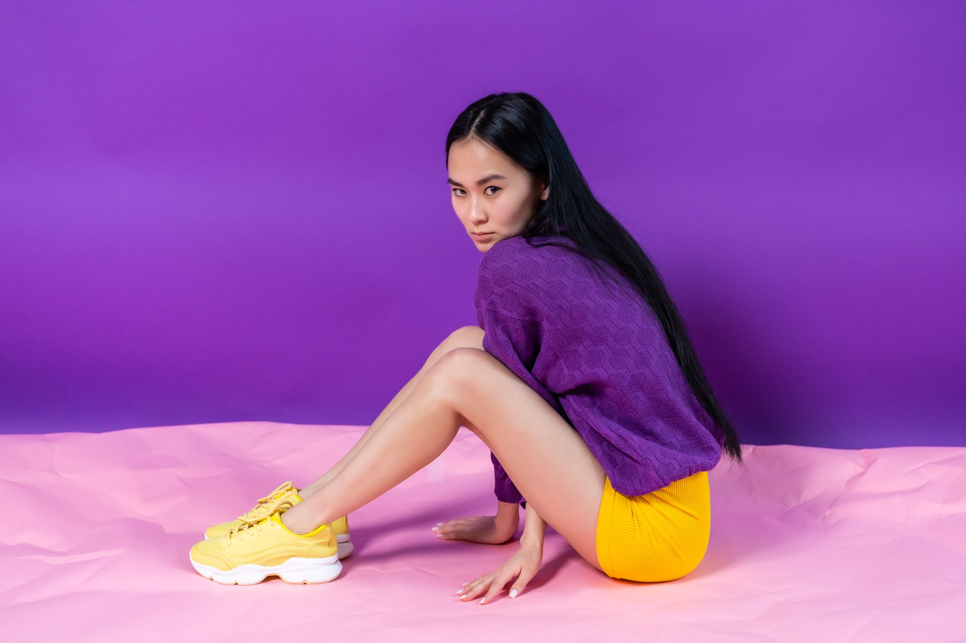 A fashionable Asian woman poses against a vivid background wearing a purple sweater and yellow shorts.